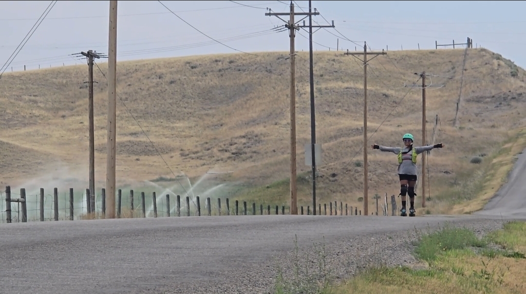 A woman roller skates on an open road, rolling hills and telephone poles in the background, her arms are wide open.